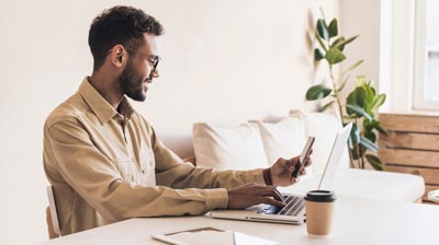 A man sitting at a desk while typing on a laptop with one hand and using his phone with his other hand