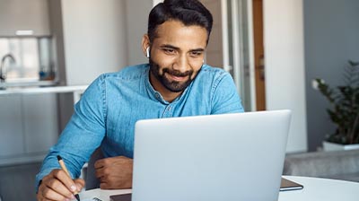 A man looking at his laptop screen while writing something down in a notebook 