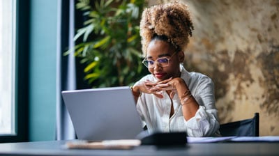 Happy African-American woman reading business report on her laptop while sitting at restaurant desk.