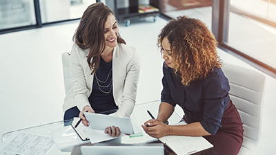 Two women colleagues dressed in business attire sitting at table conversing over a tablet.