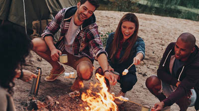 Group of young people in casual wear roasting marshmallows over a campfire while resting near the lake.