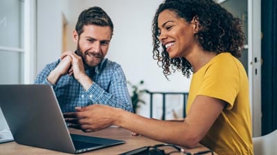 A man and a woman sitting at table while the woman is showing him something on a laptop