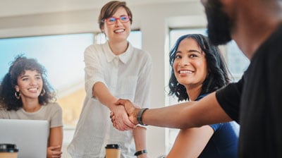 A woman standing in the center of the image between two other women sitting at a table, shakes hands with a man partially in frame to the right of the image during a meeting framed by windows in the background.