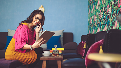 Woman in casual attire sitting in a coffee shop looking at a tablet and talking on a cellphone.