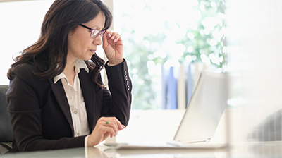 Woman dressed in business attire sitting at desk using a laptop while holding the corner of her glasses.