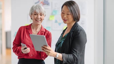 Caucasian senior businesswoman with Korean mature businesswoman standing and talking about ideas while looking at tablet computer with charts diagrams well-dressed businesswear.