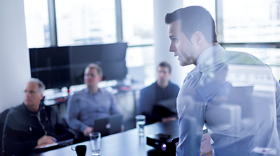 Man in business attire giving a presentation to 3 men sitting in boardroom with laptops and tablets in their hands